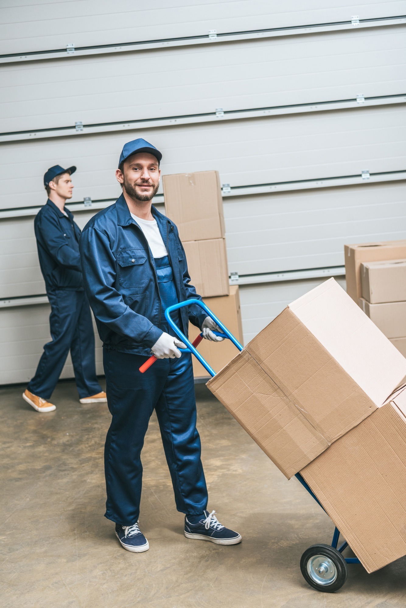 two movers in uniform transporting cardboard boxes with hand truck in warehouse